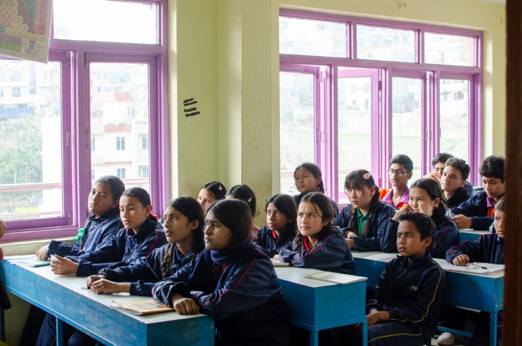 A classroom of students in dark blue uniforms sit at blue desks, attentively facing the front with notebooks open, near large purple-framed windows, eager to learn about climate and environmental stewardship.
