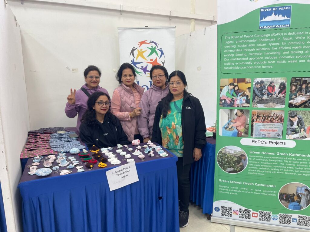 Five women stand behind a table displaying handmade crafts next to a banner explaining the River of Peace Campaign and GPF Nepal's commitment to environmental stewardship in Kathmandu.