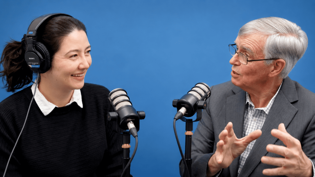 A woman and an older man sit in front of microphones, having a conversation for the Global Peace Foundation Podcast. The woman is wearing headphones and smiling, set against a solid blue background.