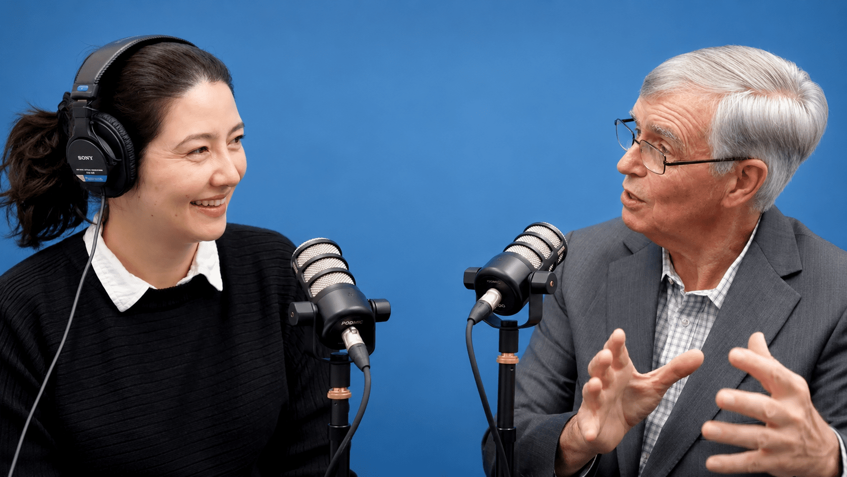 A woman and an older man sit in front of microphones, having a conversation for the Global Peace Foundation Podcast. The woman is wearing headphones and smiling, set against a solid blue background.
