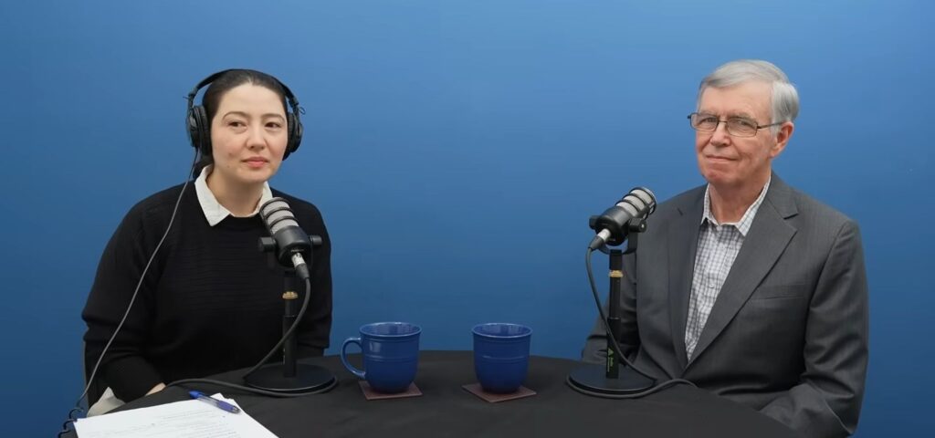 Two people sit at a table with microphones, wearing headphones, facing the camera. Between them are two blue mugs and a discussion on Sustainable Peace inspired by the Global Peace Foundation unfolds against a plain blue background.