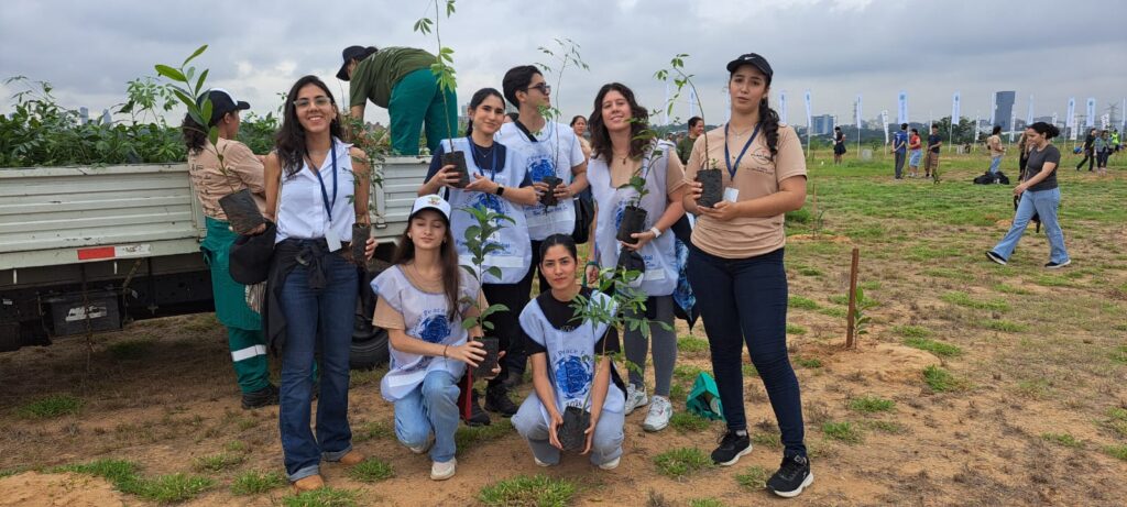 A group of people, some holding small potted plants, stand and kneel on grassy ground near a truck during the Plantatón 2026 event by Global Peace Foundation Paraguay, showcasing community-led environmental action. Other participants are visible in the background.